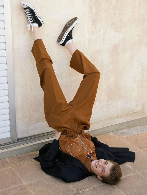 Person in upside down pose wearing brown pants, black sneakers and jacket against off-white wall and tile floor; indoor setting.