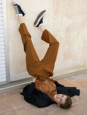 Person in upside down pose wearing brown pants, black sneakers and jacket against off-white wall and tile floor; indoor setting.