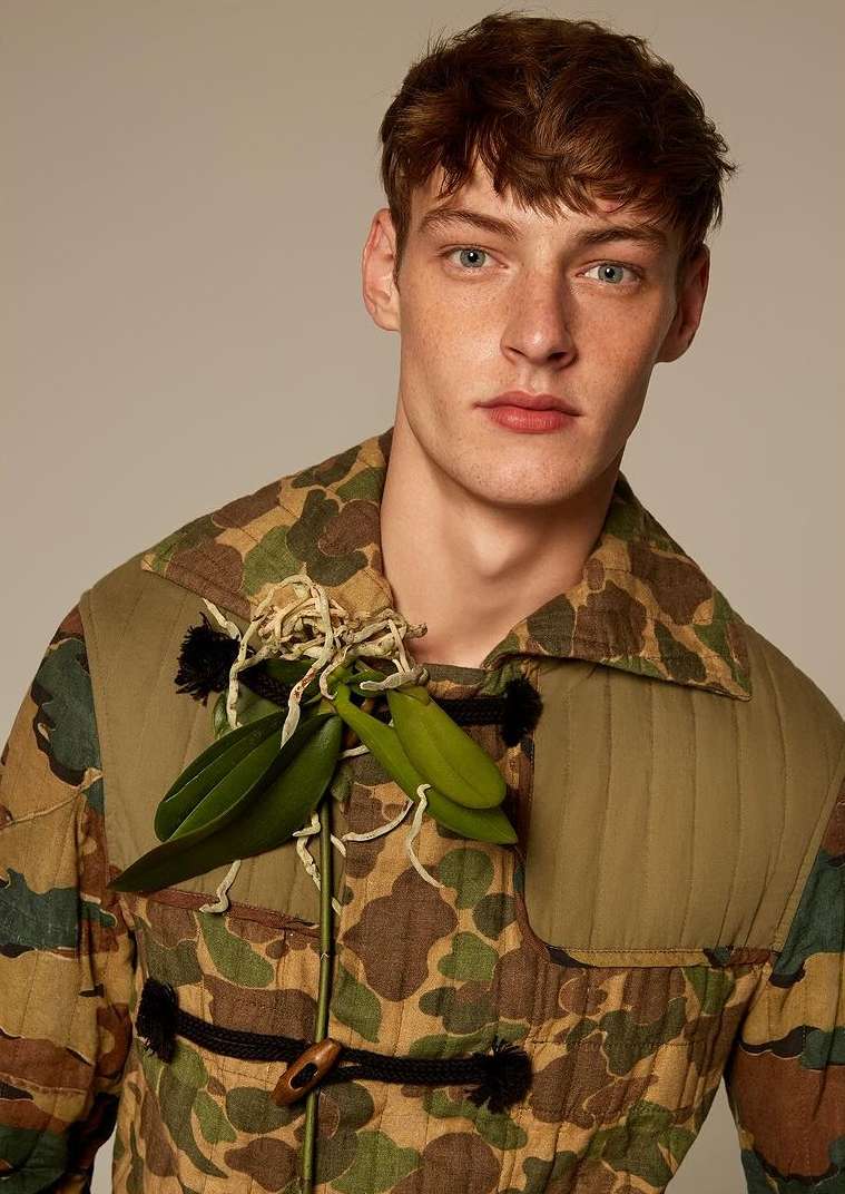 Portrait of a redhead male fashion model wearing a camo patterned jacket and decorative orchid, posing against a neutral backdrop.