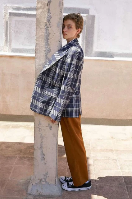 Woman in a blue and white plaid blazer standing near a weathered column in a sunlit, outdoor setting, showcasing a relaxed fashion style.