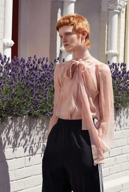 A red-haired model wearing a pink, semi-sheer blouse and black pants poses in front of a white brick wall and lavender bushes.