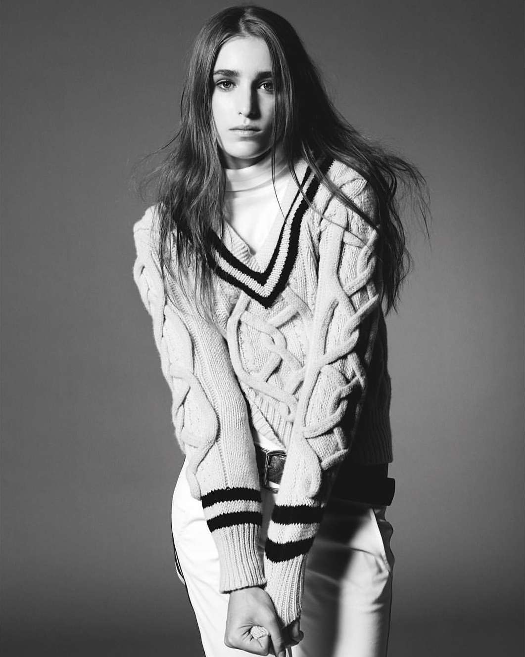 Monochrome portrait of a young adult female with long hair, wearing a cable-knit sweater, white pants, and a belt, against a gray background.
