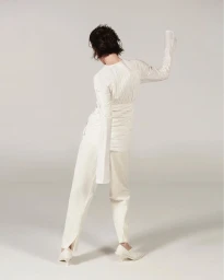 Model with dark hair wearing a white long-sleeve top and white trousers in a minimalist studio setting.