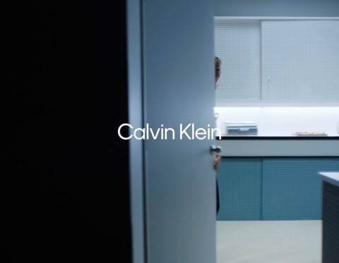 Minimalist interior scene with a person partially visible through a door, featuring the Calvin Klein logo prominently displayed.