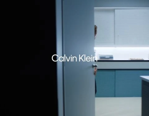 Minimalist interior scene with a person partially visible through a door, featuring the Calvin Klein logo prominently displayed.