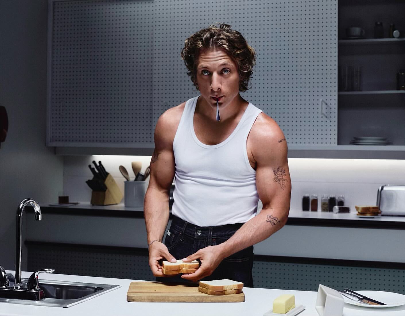 Muscular man in tank top prepares sandwich in kitchen. Cutting board, bread, and kitchenware fill the foreground in bright, soft lighting.