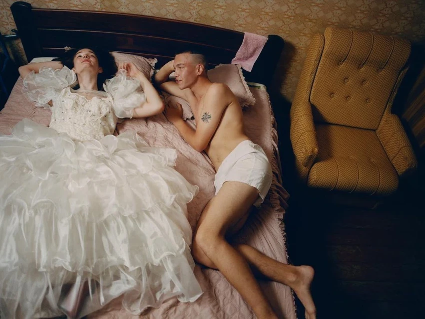Woman in white dress and shirtless man in bed with a patterned headboard, yellowish wallpaper, and yellow armchair in a domestic interior.