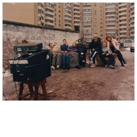 Group of people relax on a couch outdoors, in front of an old brick wall and high-rise buildings; one person wears a mask, and a vintage TV rests nearby.
