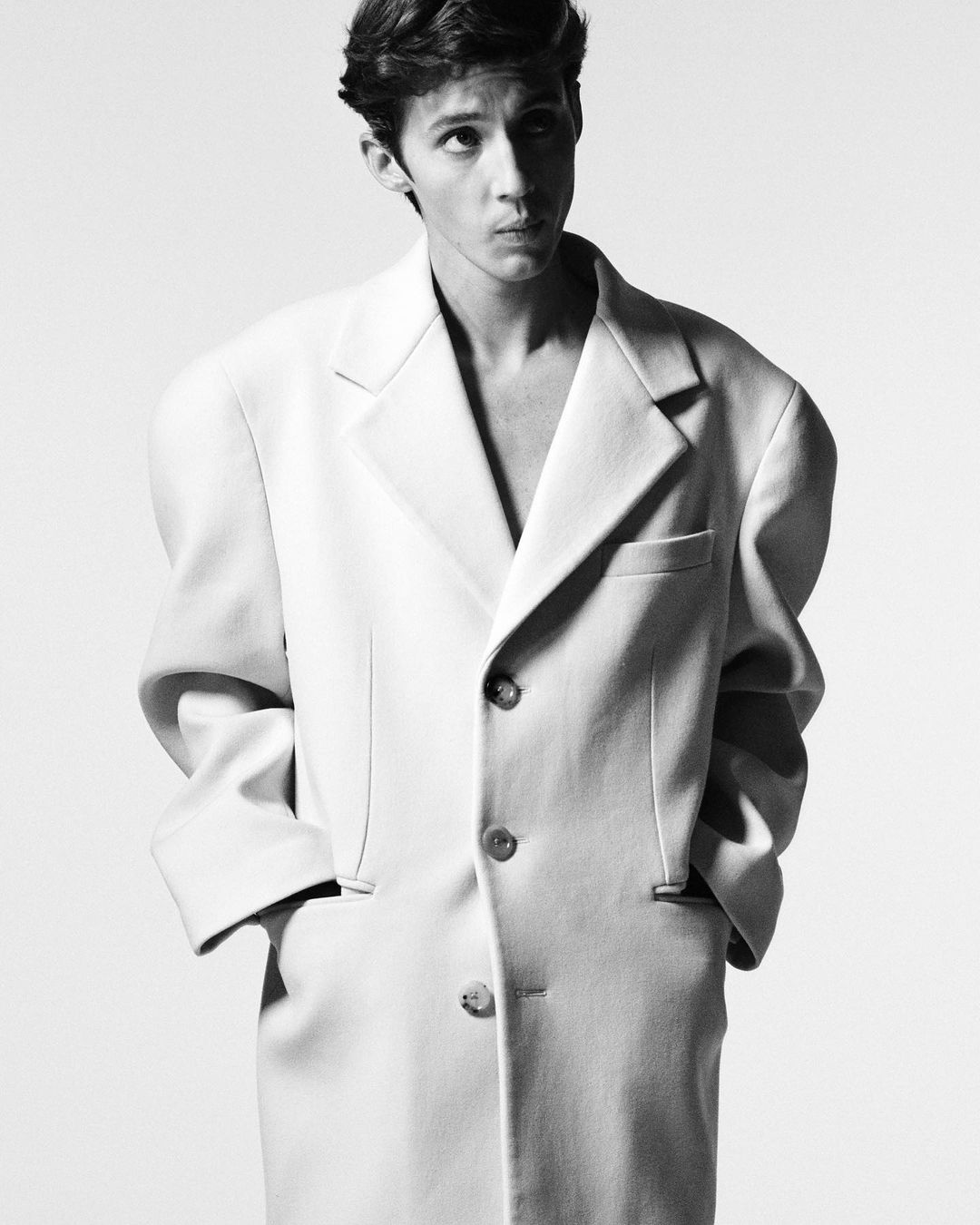 Monochrome portrait of a slender young adult male wearing a large off-white overcoat against a white background in a studio setting.