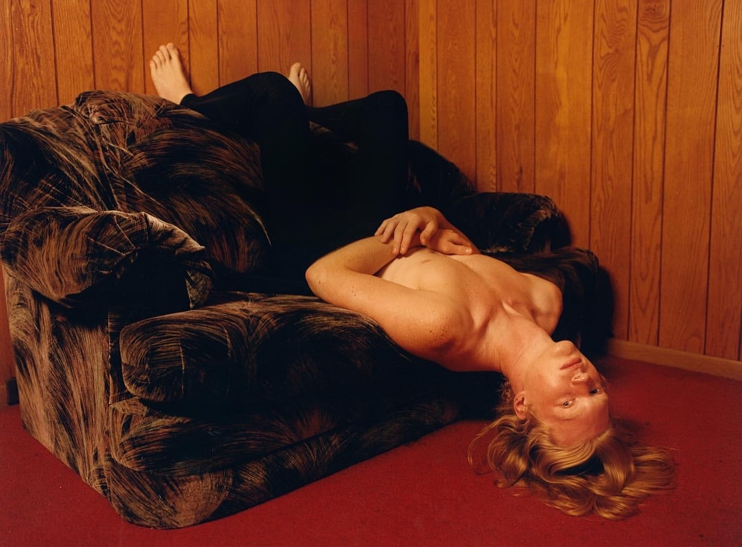 Person reclines on a dark patterned armchair in front of a wooden paneled wall, light catches the red-carpeted floor and blonde hair.