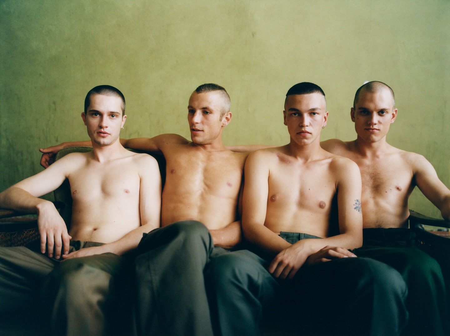 Four young adult men sit shirtless on a couch against a green wall in a medium shot with natural lighting indoors.