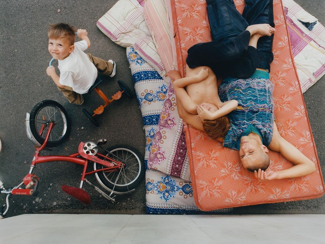 Three children outdoors with a red tricycle and patterned mattresses, viewed from overhead in bright daylight.