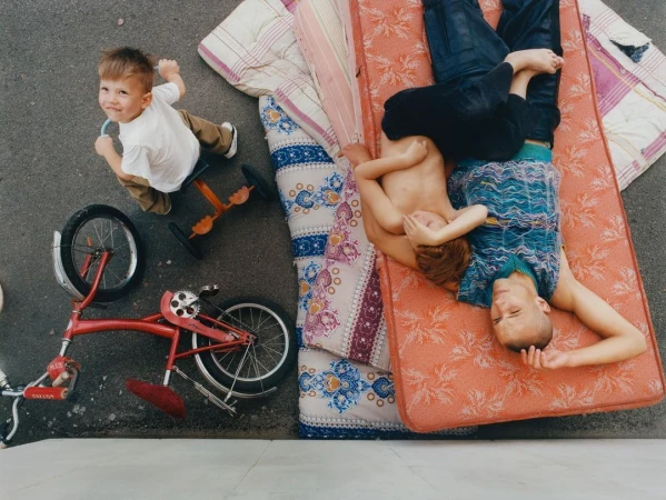 Three children outdoors with a red tricycle and patterned mattresses, viewed from overhead in bright daylight.
