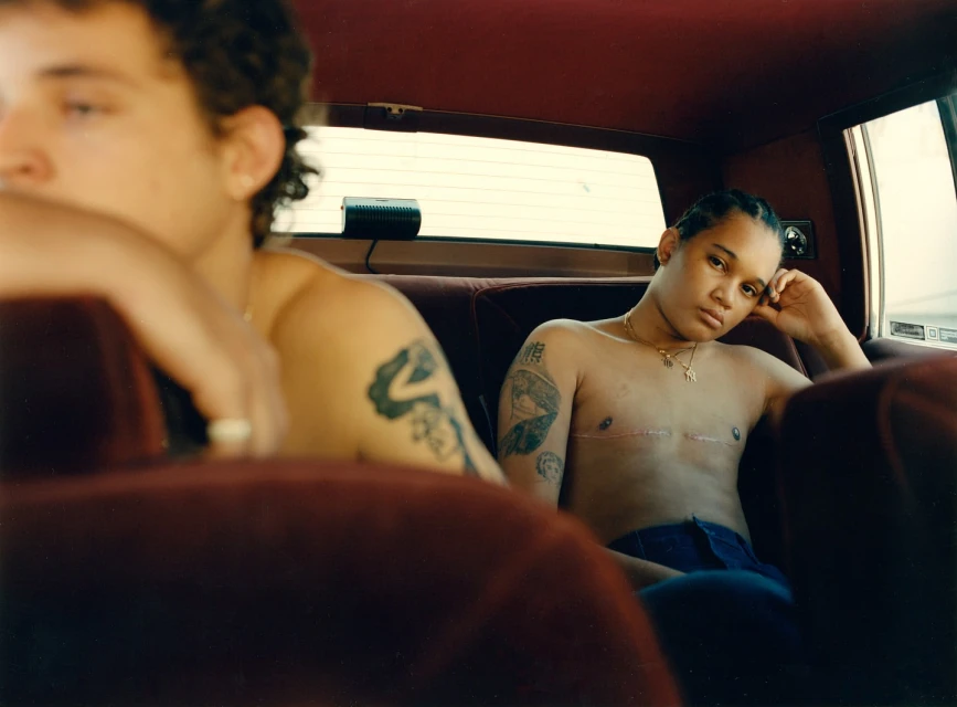 Two topless people ride in the back seat of a car. One person faces the camera with a slight smile. Tattoos are visible on their arms.