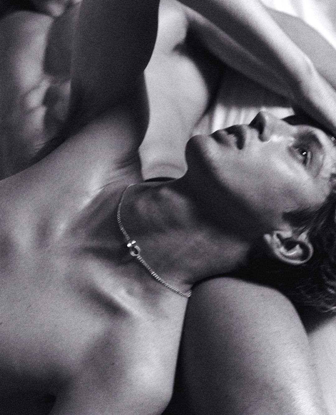 Black and white photograph of a shirtless man with a necklace lying on a white surface with another person beside him.