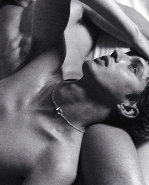 Black and white photograph of a shirtless man with a necklace lying on a white surface with another person beside him.