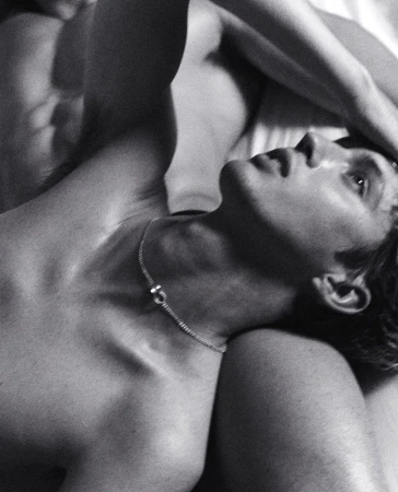 Black and white photograph of a shirtless man with a necklace lying on a white surface with another person beside him.