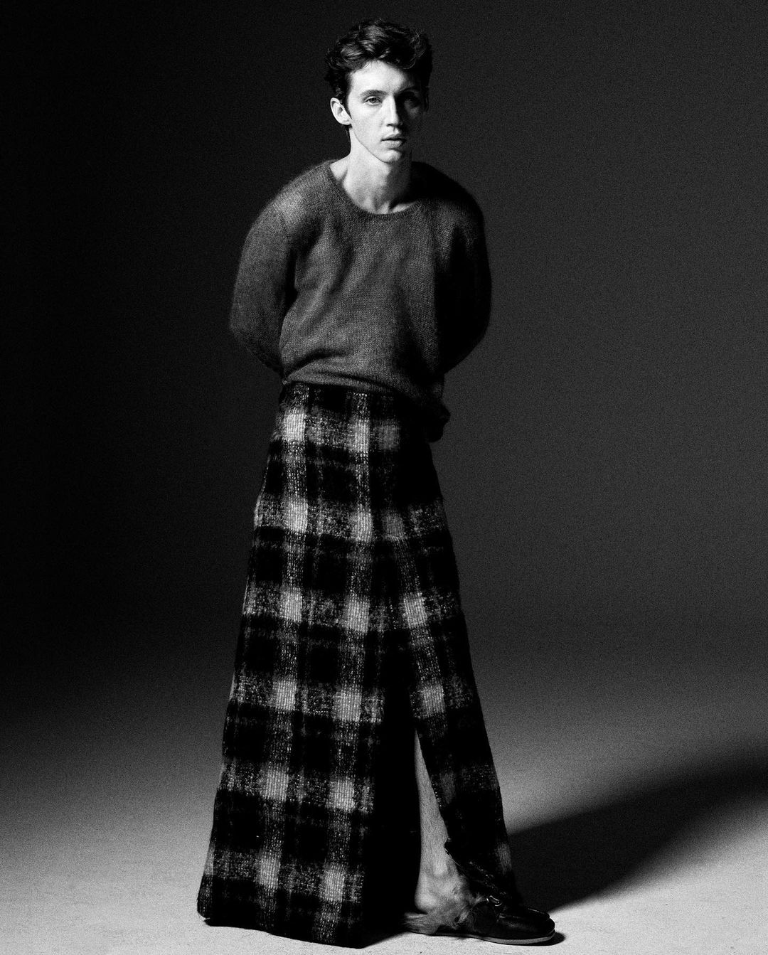 Monochrome portrait of a young adult male in a wool sweater and plaid skirt, set against a dark studio backdrop with soft, diffused lighting.