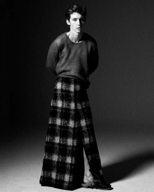 Monochrome portrait of a young adult male in a wool sweater and plaid skirt, set against a dark studio backdrop with soft, diffused lighting.