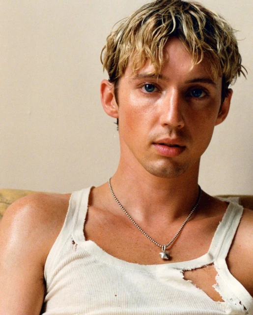 Blonde man with blue eyes poses in a white tank top. He wears a silver necklace indoors. The lighting is soft and even.