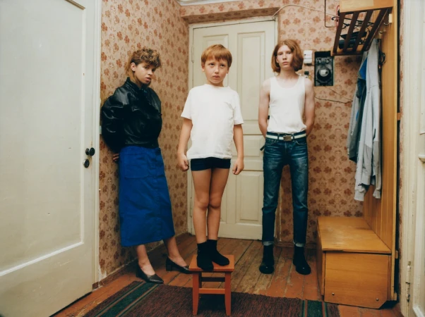 Three children pose in an indoor setting with patterned wallpaper, showcasing vintage fashion and a candid, documentary-style aesthetic.