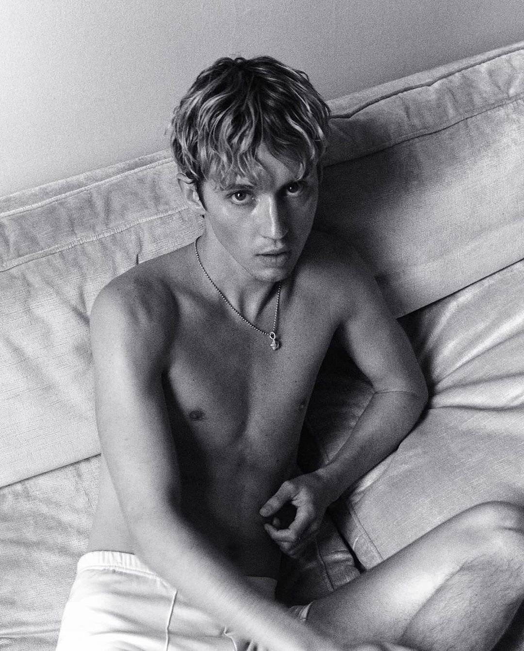 Black and white portrait of a young adult male with blond hair, sitting on a light-colored sofa in a relaxed pose indoors.