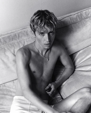 Portrait of shirtless young man with curly hair and pendant necklace seated on a sofa photographed by Stuart Winecoff