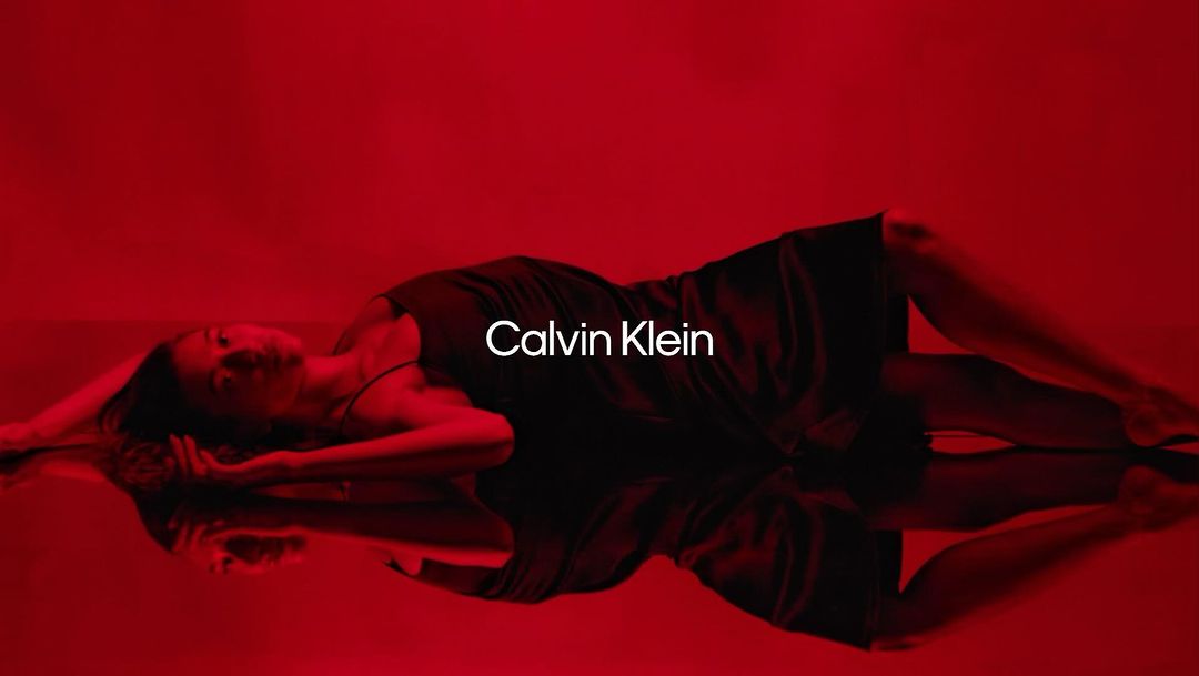 Adult woman in a black dress lies on a reflective surface, lit with red light, promoting Calvin Klein. Fashion and beauty shot.