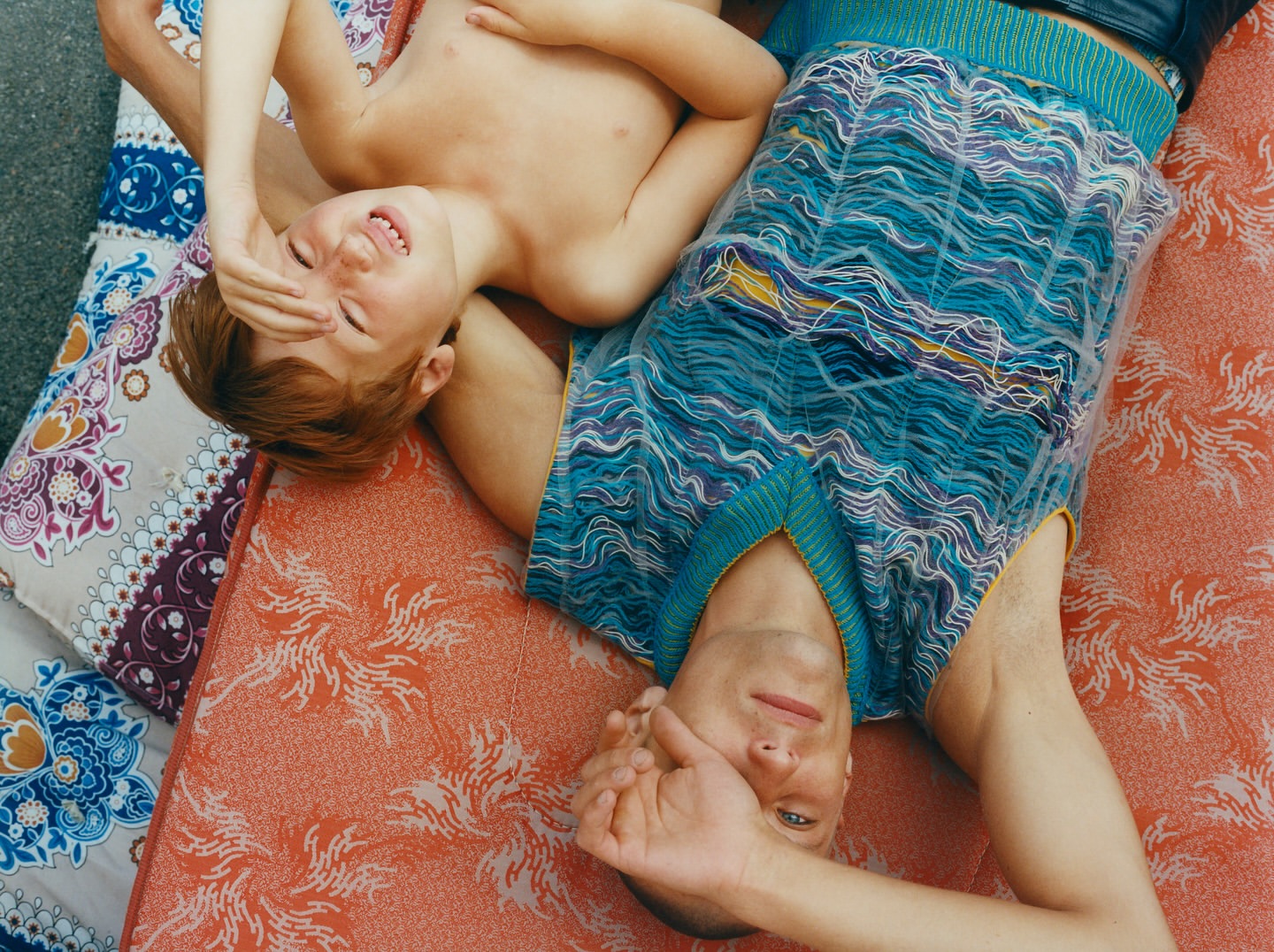 A boy and an adult male lie on patterned mattresses outdoors, covering their faces with their hands, both smiling in bright daylight.