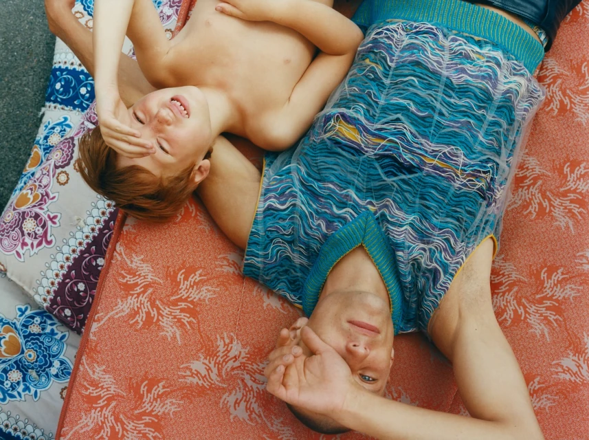 A boy and an adult male lie on patterned mattresses outdoors, covering their faces with their hands, both smiling in bright daylight.