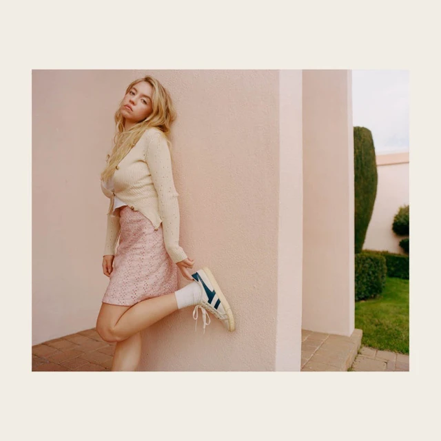 A blonde woman in a mini skirt poses against a building in a cream-colored sweater and white sneakers with blue details in an eye-level outdoor shot.