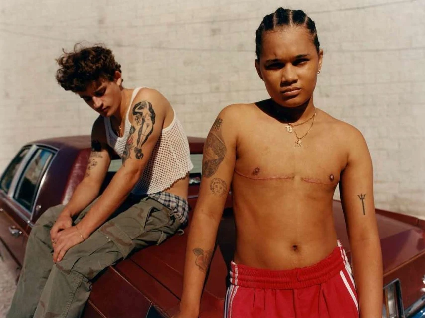 Two tattooed people pose near a maroon car, one seated on the hood, one standing, against a plain wall in outdoor daylight.