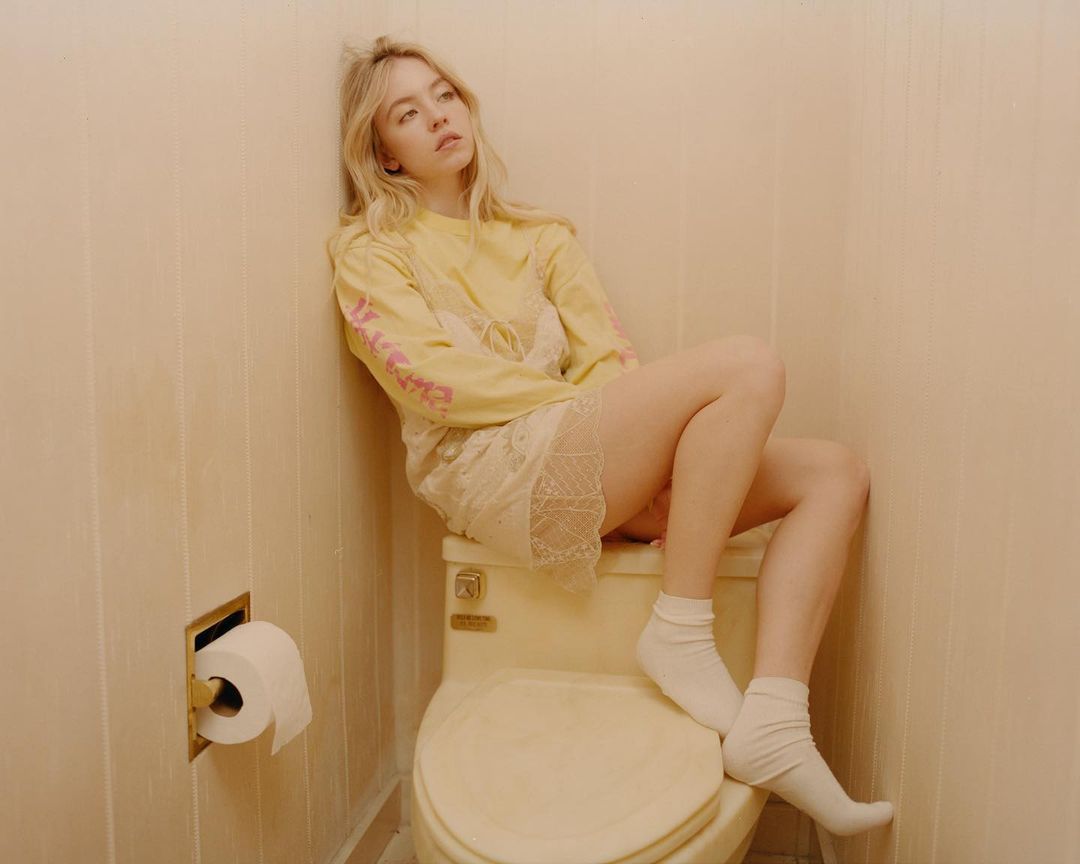 Blonde woman in a casual outfit, sits on a toilet in a bathroom with light beige walls, facing forward with a contemplative expression.