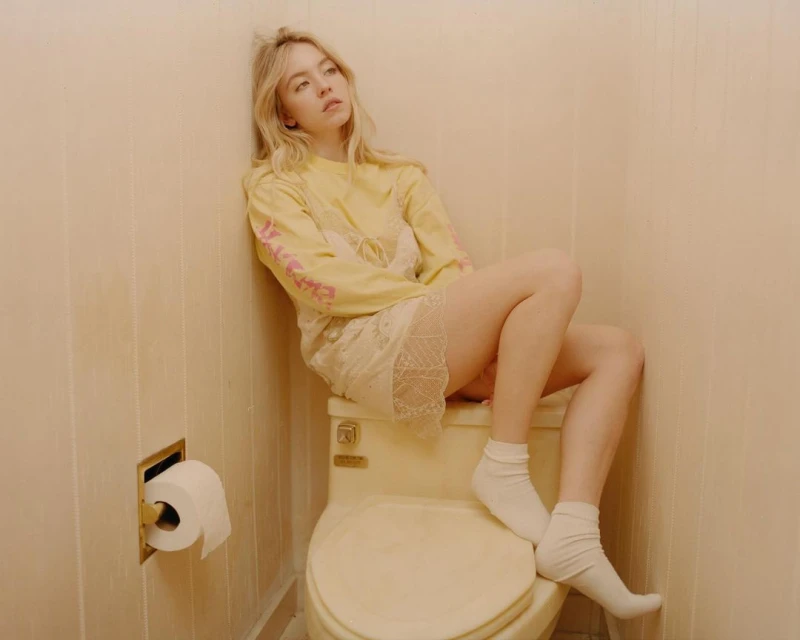 Blonde woman in a casual outfit, sits on a toilet in a bathroom with light beige walls, facing forward with a contemplative expression.