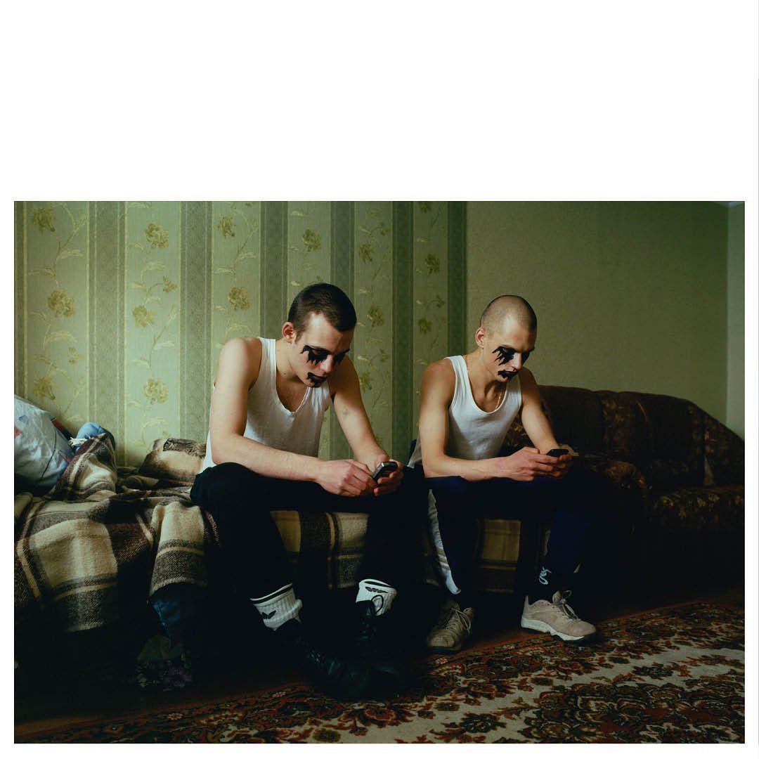Two adult males with dark eye makeup sitting on a bed and couch, each using smartphones in an indoor, residential setting.  