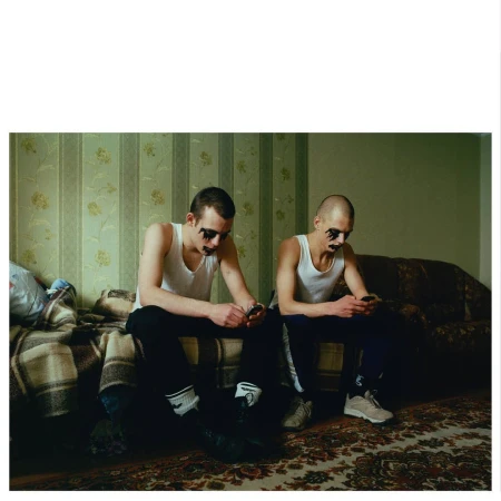 Two adult males with dark eye makeup sitting on a bed and couch, each using smartphones in an indoor, residential setting.  