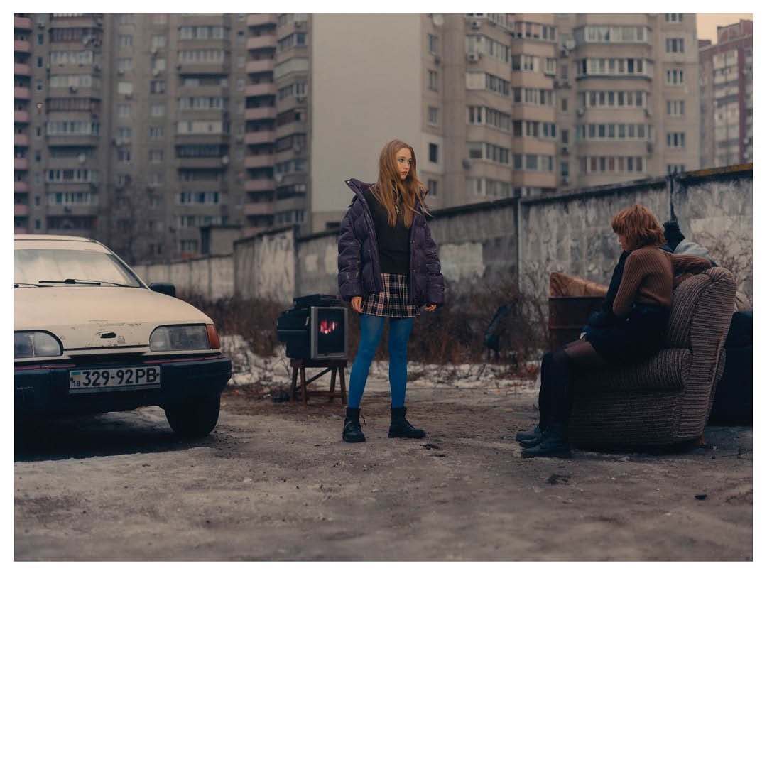 Two women in an urban setting with an old car and apartment buildings. One woman stands, the other sits outside on a weathered armchair.