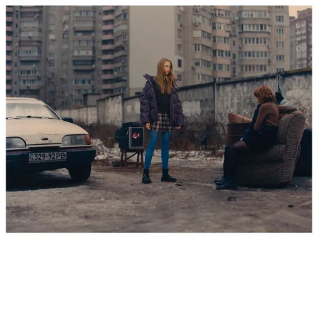 Two women in an urban setting with an old car and apartment buildings. One woman stands, the other sits outside on a weathered armchair.