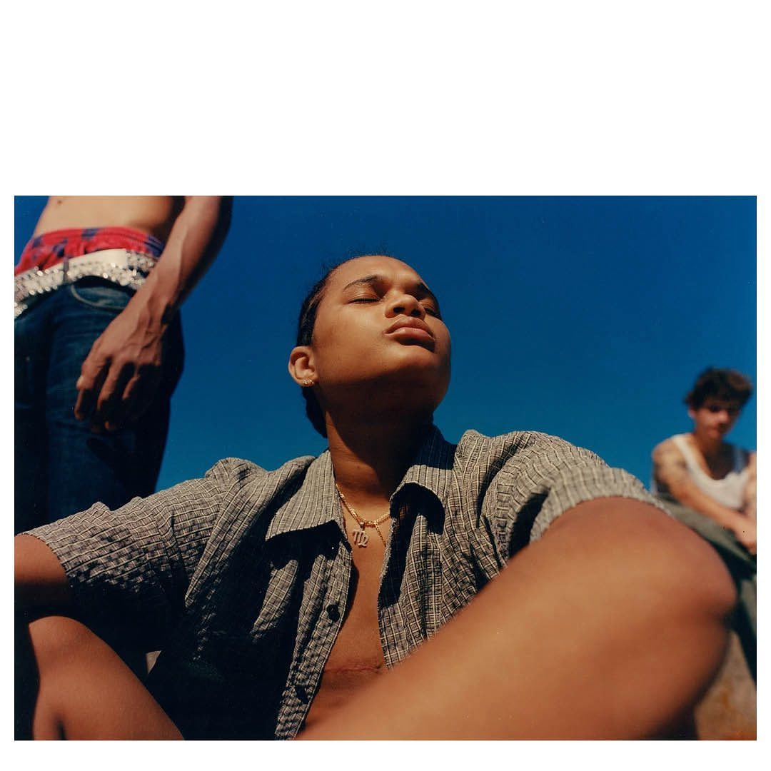 Portrait of an adult in unbuttoned shirt with gold chain against a blue sky backdrop, featuring two additional people in the background.