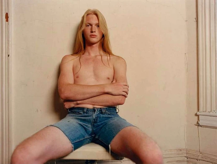 Shirtless young man with long blond hair and crossed arms wearing denim shorts sitting indoors by a white wall