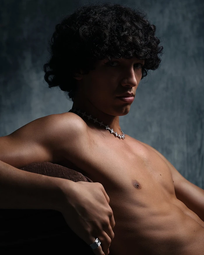 Portrait of a shirtless young man with dark curly hair and a silver chain necklace looking seriously toward the camera in Paris.