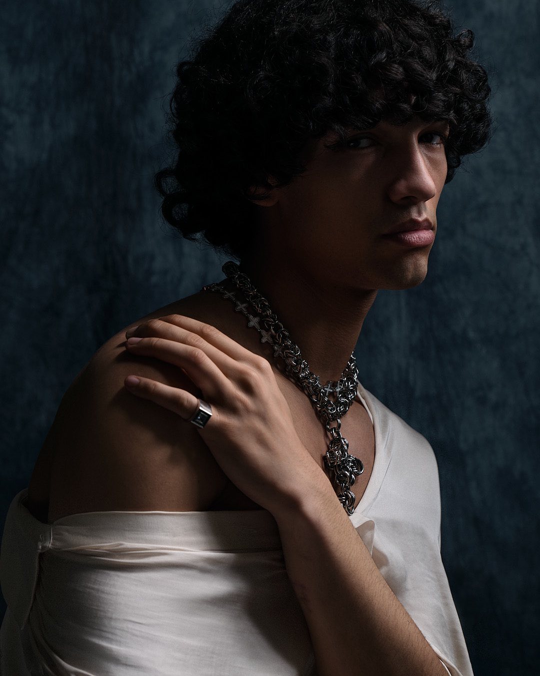Close-up portrait of a man with curly dark hair wearing a white garment and an ornate silver chain necklace, against a textured blue backdrop.