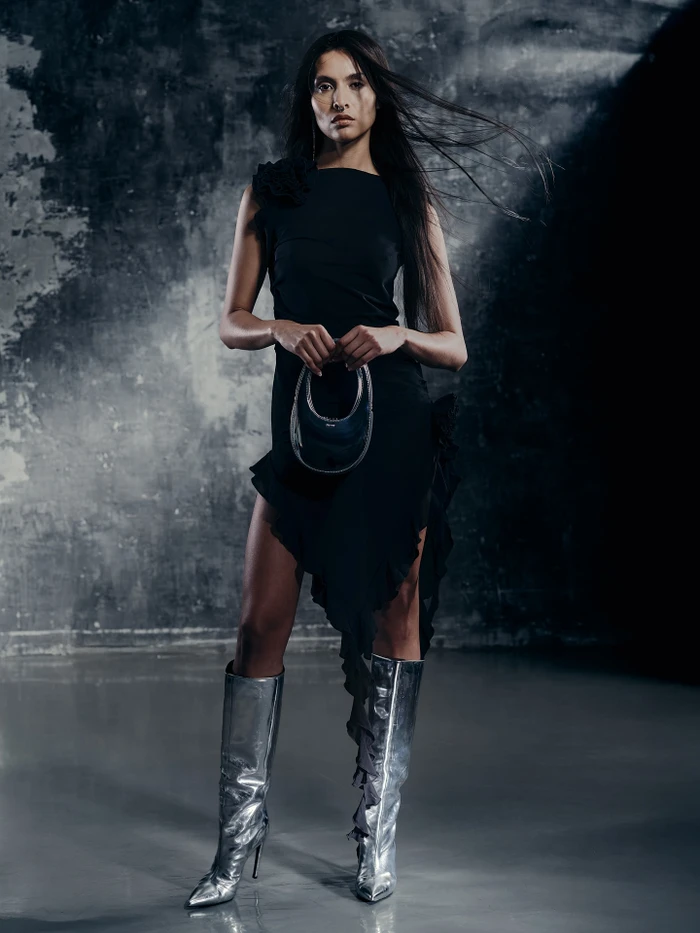 Woman wearing a black dress and metallic silver boots holding a handbag against a textured backdrop