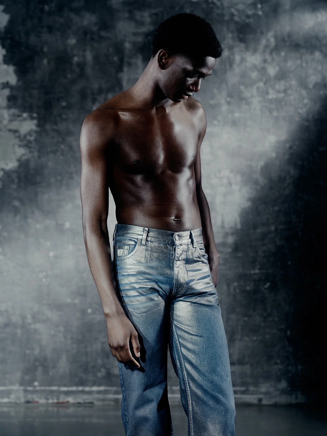Shirtless black man wearing shiny metallic silver denim jeans against a textured grey wall in a studio portrait by Caroline Pusset