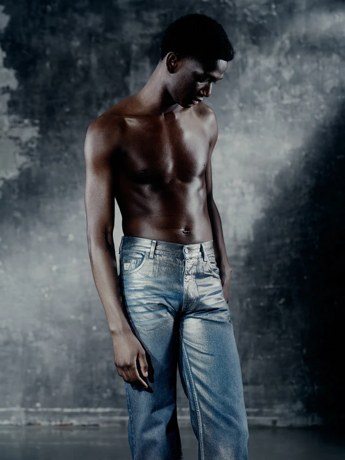 Shirtless black man wearing shiny metallic silver denim jeans against a textured grey wall in a studio portrait by Caroline Pusset