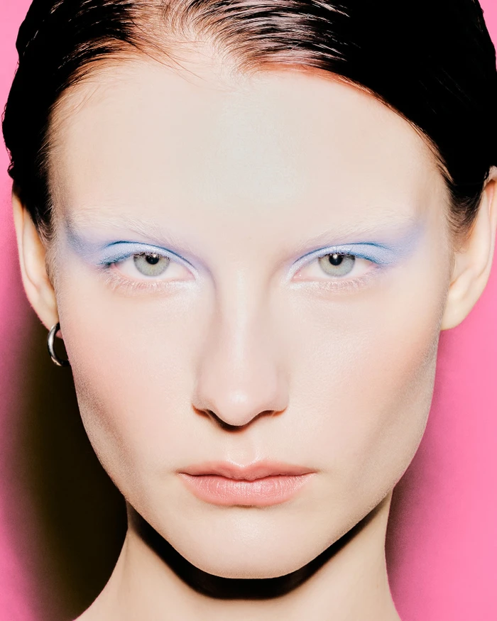 Close-up portrait of a model with light blue eyeshadow, bleached eyebrows, and slicked dark hair against a pink background.