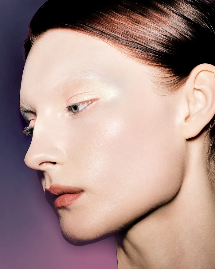 Close-up profile portrait of a woman with highlighted cheekbones and glossy nude lips against a gradient purple background.