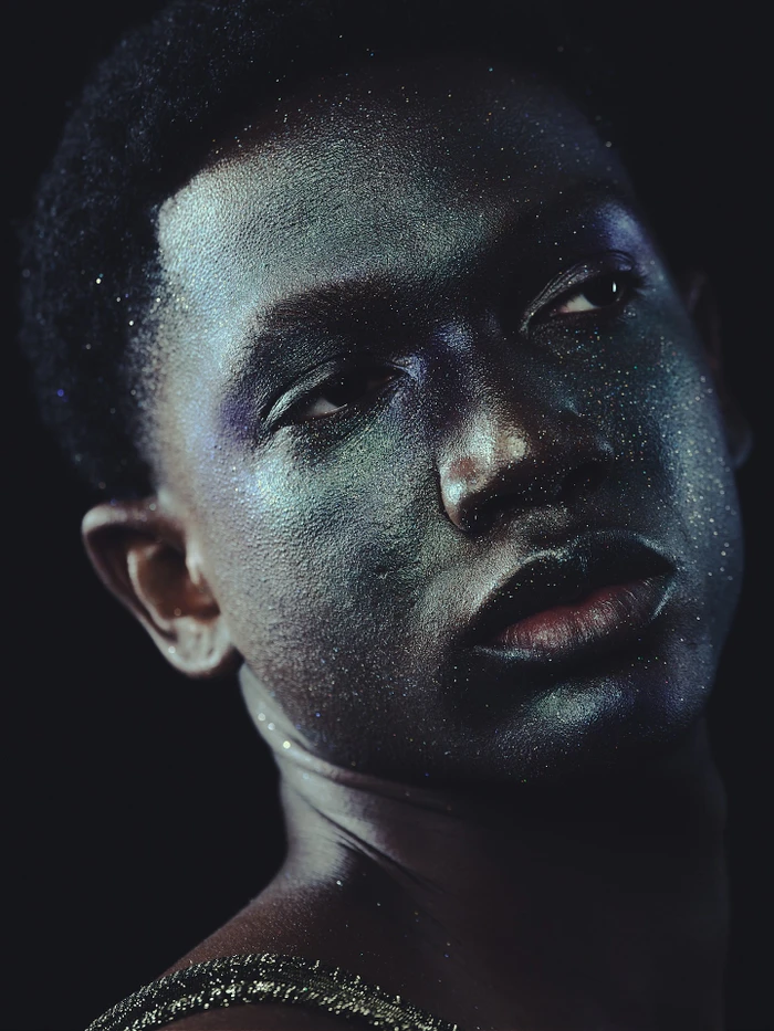 Close-up portrait of a Black man with metallic green glitter makeup on his face and shoulders against a dark background.