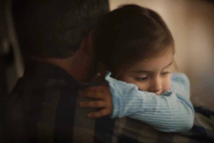 Close-up portrait of a young child in a blue ribbed top resting her head on a man's shoulder