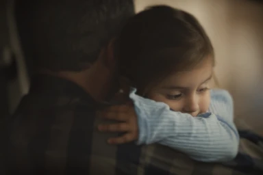 Close-up portrait of a young child in a blue ribbed top resting her head on a man's shoulder